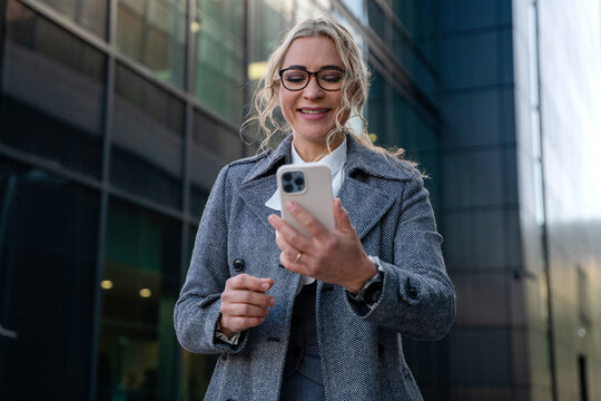 Woman smiles while looking at smartphone outside a modern office building during the daytime
