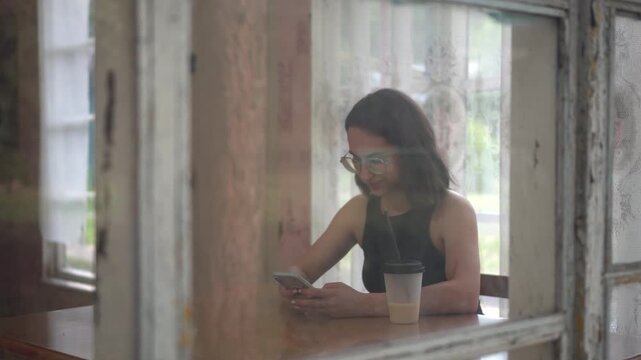 Young woman using smartphone and drinking coffee while sitting on old glass veranda