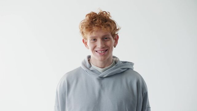 Surprised young Caucasian redhead man opening mouth in excitement and looking at camera in bright studio. Casual male wearing gray hoodie and expressing genuine amazement with copy space.