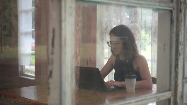 Young woman working on laptop and drinking coffee at table on old glass veranda with panoramic camera movement
