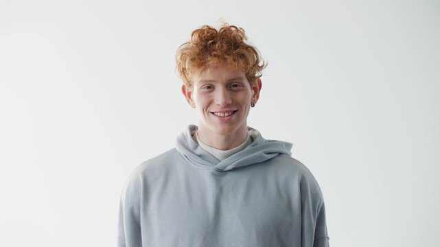Happy young Caucasian redhead man smiling warmly at camera in bright studio. Casual male wearing gray hoodie and expressing genuine happiness. Friendly positive portrait with copy space.