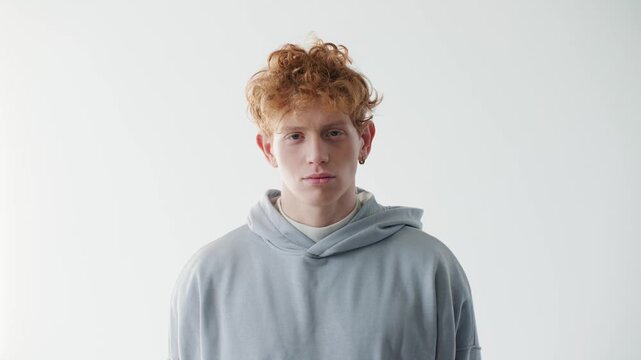 Serious young Caucasian redhead man standing and looking at camera in bright studio. Casual male wearing gray hoodie and showing calm focused expression. Confidence and determination with copy space.