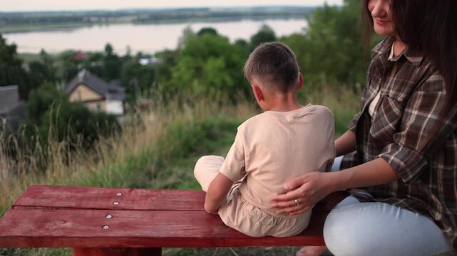 Close up slow motion shows mother gently comforting son on hilltop bench. Beautiful lake village forest and fields surround loving family moment with parental support.