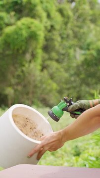Vertical video. Woman Rinses an Empty White Plant Pot with a Garden Hose, Cleaning the Container Outdoors before Reuse for Sustainable Gardening