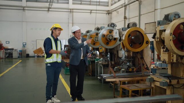 Two men walk through a factory floor. One wears a suit, the other a safety vest. They gesture toward machines and discuss operations