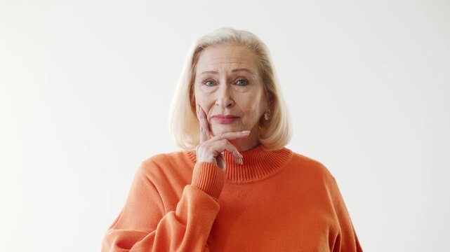 Senior Caucasian woman standing and looking at camera with confident calm expression. Elegant elderly female wearing orange sweater. Clean studio background with copy space for design.