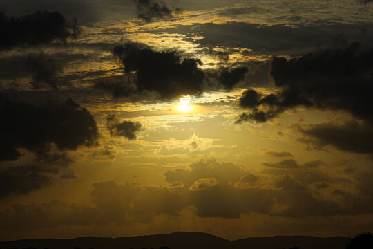 Sunset glow illuminates layered clouds hanging low over a silhouette of rolling hills in the distance.