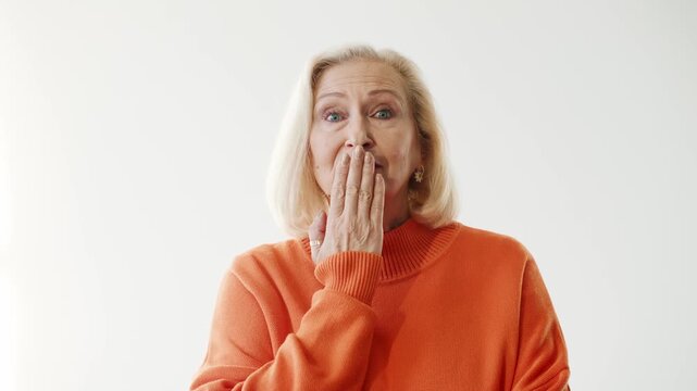 Surprised senior Caucasian woman covering mouth with hand while looking at camera. Elderly female expressing shock disbelief or unexpected news. Bright studio background copy space.