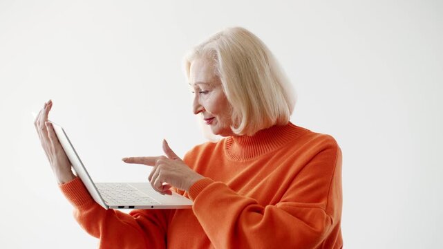 Elegant senior Caucasian woman using laptop while standing against clean background. Female browsing online working remotely or learning new skills. Modern technology active lifestyle.