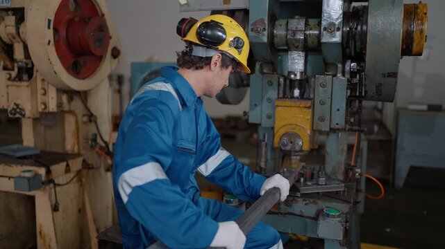 Worker wears hard hat, blue jumpsuit, and white gloves. He operates heavy industrial press with focused expression