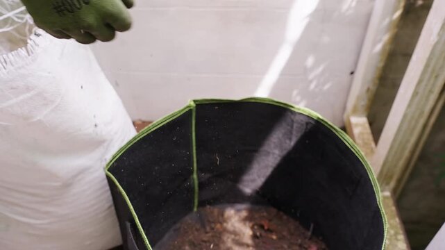 Gardener Wearing Protective Gloves Transfers Dark Soil from a White Bag into a Black Fabric Pot, Preparing Substrate for Container Planting