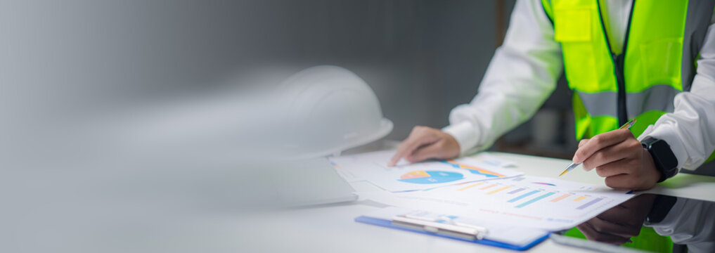 engineer writing and reviewing financial graph chart document on table. Male construction worker analyzing building project data report, safety vest and hardhat background concept.