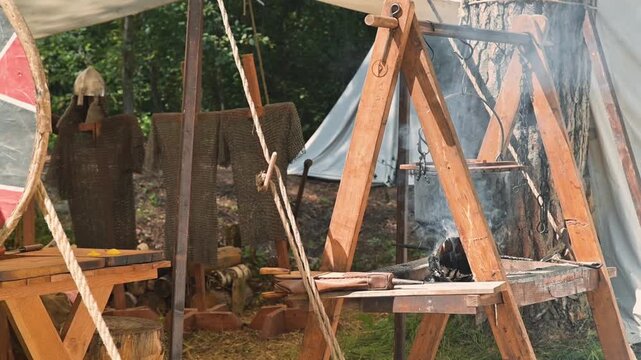 A medieval reenactment area with wooden frame structures and smoke rising from a fire. Individuals dressed in simple cloaks engage in various activities, reflecting life in the early Middle Ages