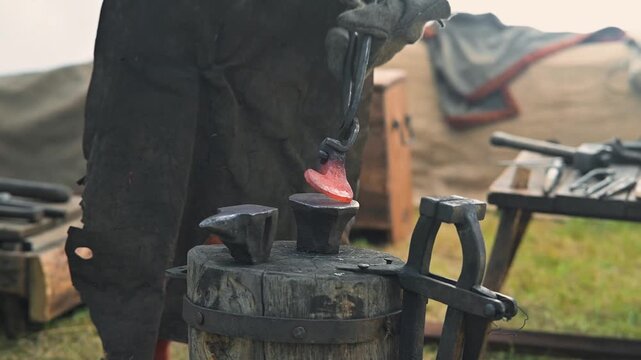 In a historical reenactment, a blacksmith works at an anvil, heating metal in a forge. The blacksmith wears traditional clothing, focusing on crafting tools in a rustic outdoor setting