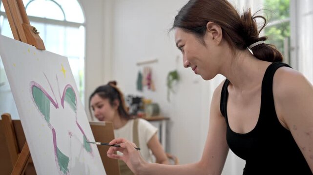 Two young Asian women painting a colorful landscape on canvas together during a relaxing weekend at home, enjoying creative art as a hobby in a bright living room