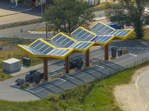 Aerial view of an electric vehicle charging station featuring a distinctive yellow zig-zag roof with integrated solar panels, with cars parked at charging units in the Netherlands.