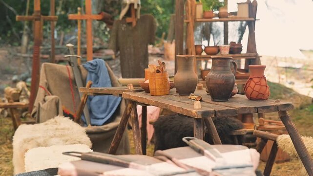 A medieval setting shows a wooden table with pottery and tools arranged neatly. Various handmade items are visible alongside simple fabrics and natural surroundings