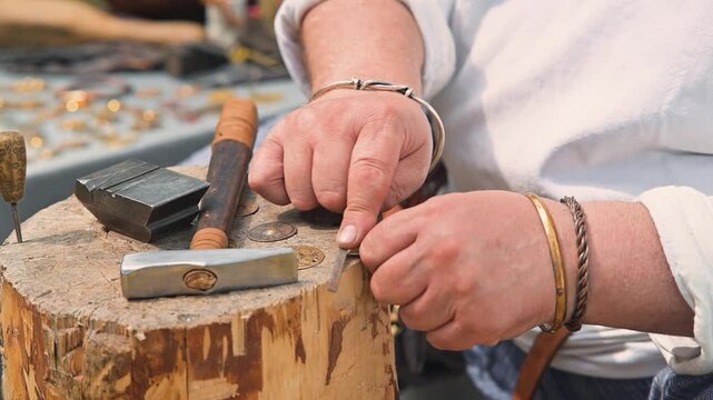 Hands are busy shaping metal on a wooden stump using a hammer and other tools in a medieval workshop. The setting shows a rustic atmosphere with various tools scattered on a table nearby