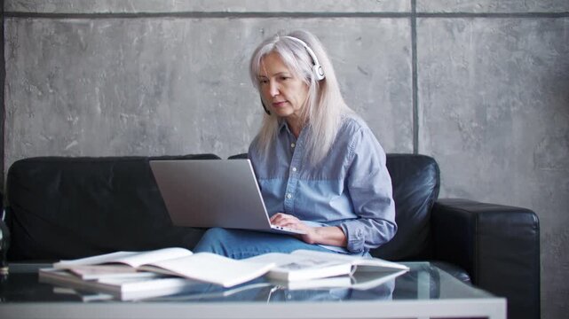 Senior Caucasian woman wearing headset and using laptop during online meeting from home office. Mature female talking with clients and working remotely from comfortable sofa. Distance communication.