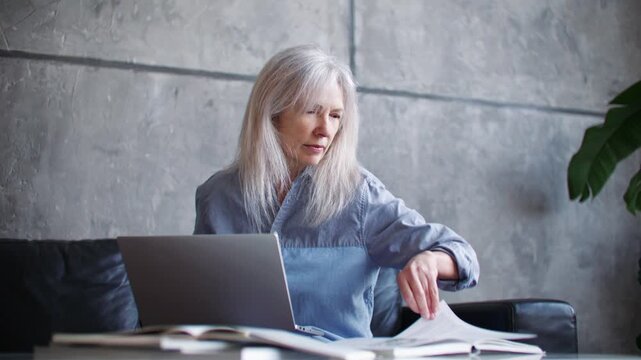 Senior Caucasian woman working on laptop and looking away while thinking at home office. Mature female reviewing documents and planning tasks at desk. Remote work. Business lifestyle.