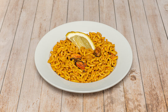 Spanish fideu&agrave; noodle dish with mussels and lemon wedge, served on white plate over light wood table