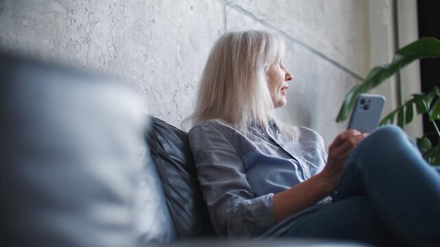 Happy mature Caucasian woman using smartphone while resting on comfortable sofa at home. Senior female reading messages and smiling at mobile screen. Cozy living room. Modern technology.