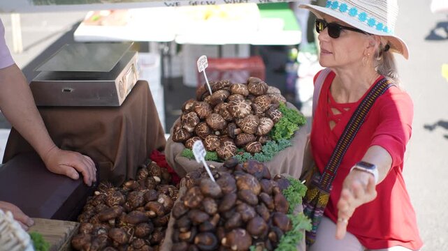A smiling woman selects fresh produce and mushrooms at the Sonoma Farmers Market on a sunny morning while enjoying her time.