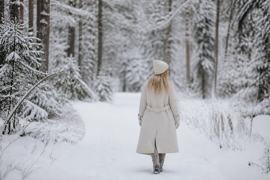 A woman in a coat and beanie walks down a snowy, wooded path during winter.