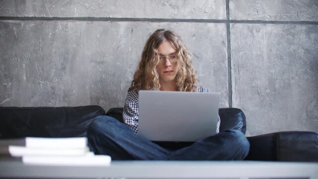 Long haired Caucasian man wearing eyeglasses using laptop while sitting on sofa in modern home office. Male working remotely and focusing on online project. Comfortable workspace indoors.