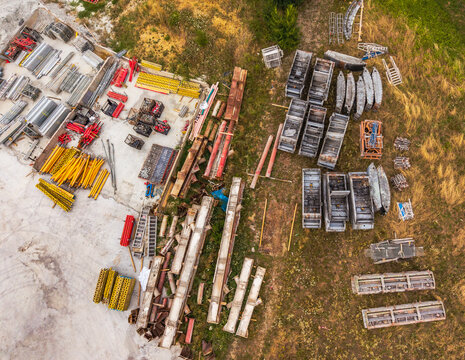 Aerial view of a construction site with stacks of metal scaffolding, yellow support beams, and building materials arranged on concrete and grassy ground in Serravalle Scrivia, Piedmont, Italy.