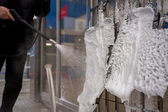 Man spraying water with high pressure washer gun onto black EVA car floor mat hanging on rack at self service car wash station outdoors.