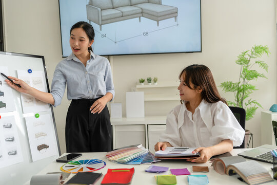 Two Asian women working together in a bright, modern studio.while using a laptop and smartphone. The creative workspace is filled with colorful samples for seasonal palette selection.