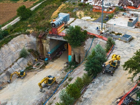 Aerial view of the Serravalle construction site featuring a tunnel entrance, yellow excavators, a mobile crane, and industrial equipment in Serravalle Scrivia, Piedmont, Italy.