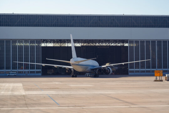White airplane rests inside a vast industrial hangar at the airport for repairs while a small vehicle drives across the sunny concrete tarmac nearby