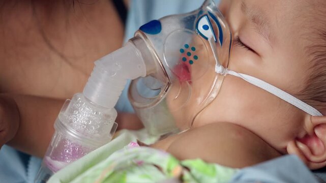Sleeping infant boy receives respiratory nebulizer therapy while being held by a parent, illustrating pediatric hospital care for breathing problems, illness, infection, and recovery.