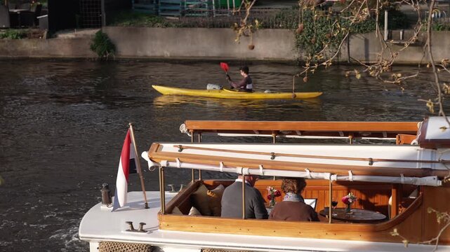 A high-angle view of a couple enjoying a private luxury boat cruise on an Amsterdam canal, complete with fine dining setup and a flying Dutch flag, while a kayaker paddling a yellow kayak