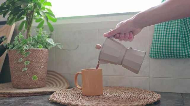 unrecognizable person's hand pouring hot coffee from a traditional italian moka pot into a ceramic mug. The cozy kitchen scene in a  moment of morning wellbeing