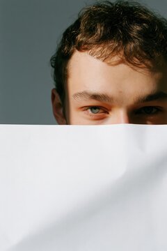 Young man peeking over a white paper sheet on a gray background for minimal banner and poster design, subtle studio portrait for modern background and creative layout