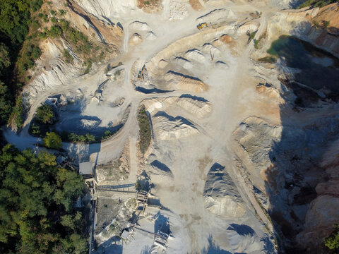 Aerial drone view of an active open-pit quarry with terraced terrain. Unpaved haul roads and stockpiles of extracted aggregate in a mining pit. Machines and excavators in industrial sand quarry.