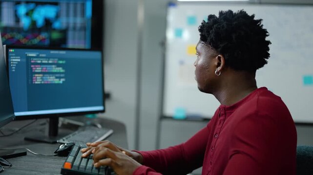 Black male software developer typing code on computer in dark office. Focused African American programmer working on software engineering, web development, data analysis, and technology project.