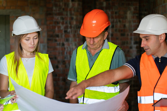 Builder points to fragment of floor plan standing near colleagues. Construction worker tells architects and partner about specifics of layout