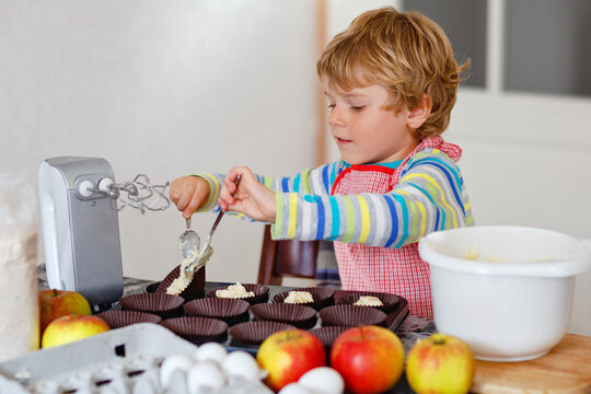 Cute little happy blond preschool kid boy baking apple cake and muffins in domestic kitchen. Funny lovely healthy child having fun with working with mixer, flour, eggs, fruits. Little helper indoors