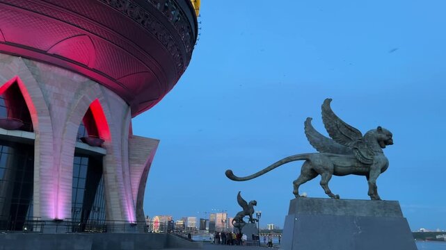 Low-angle twilight view of illuminated observation tower crown beside river, with griffin sculptures, ornate arches, circular symmetry and decorative metalwork against cool urban skyline
