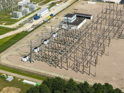 Aerial view of an electrical substation with high-voltage power lines, metal structures, and transformers set on a gravel ground surrounded by green grass and trees in the Netherlands.