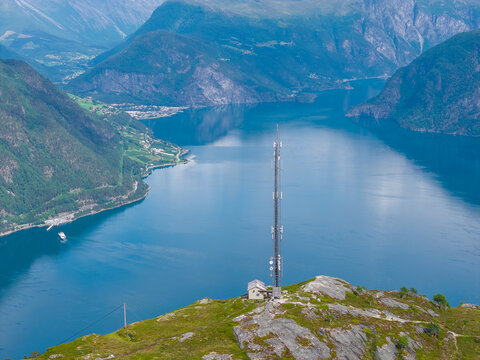 Aerial view of a tall communication tower standing on a rocky hilltop overlooking the deep blue waters of a fjord surrounded by steep mountains in Sylte, More og Romsdal, Norway.