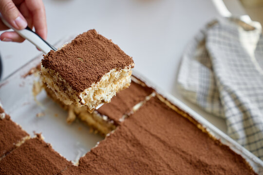Hand lifts tiramisu portion from glass dish, with cocoa topping and cream layers visible