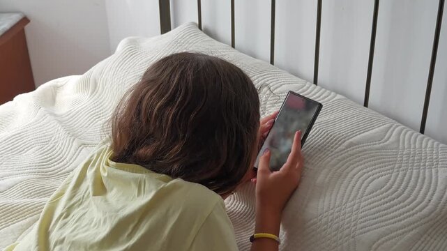 Top down view of a teenage girl with brown hair lying on the bed, intently scrolling and swiping left with her finger through content on her mobile phone