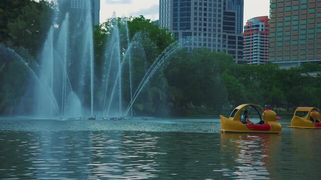 Lumpini Park fountain and pedal boats on lake with Bangkok city skyline, People riding duck boats on water among skyscrapers in Thailand, slow motion 4k