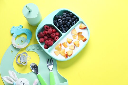 Baby tableware. Section plate with food, bottle, cutlery, silicone bib, toys and pacifier on yellow background, flat lay. Space for text