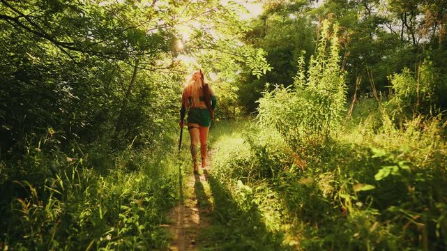 Fantasy elf fairy tale woman amazon hunts waks in forest green trees grass sun light flare backlit, sexy girl hunter holds long bow in hands back rear view. art video creative costume long blonde hair
